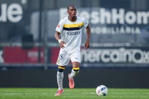 Soccer player in a white kit with yellow trim dribbling a ball on a green pitch with stadium boards in the background.