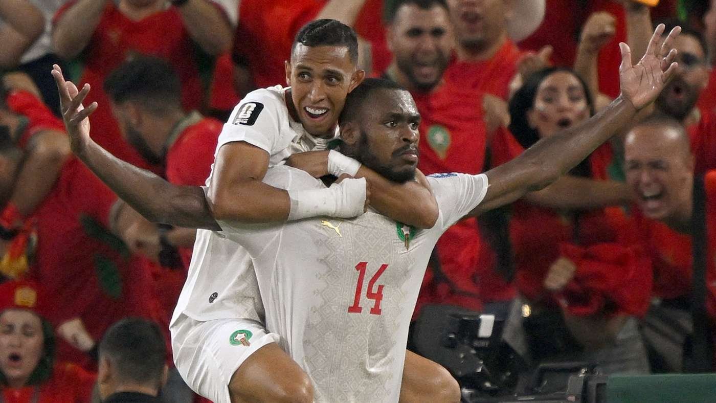 Two soccer players celebrate a goal; one lifts the other on his shoulders as red-clad fans cheer in the background.