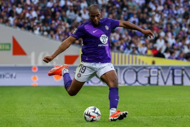 Soccer player in a purple jersey kicking a ball during a match, number 19 visible on shorts, stadium crowd in background.