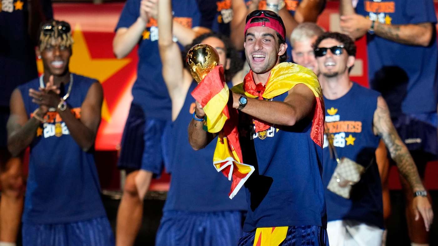 Joyful athlete in a red cap holds a gold trophy while wrapped in a red-yellow Spanish flag, surrounded by teammates in blue uniforms celebrating. Joyful athlete in a red cap holds a gold trophy while wrapped in a red-yellow Spanish flag, surrounded by teammates in blue uniforms celebrating.