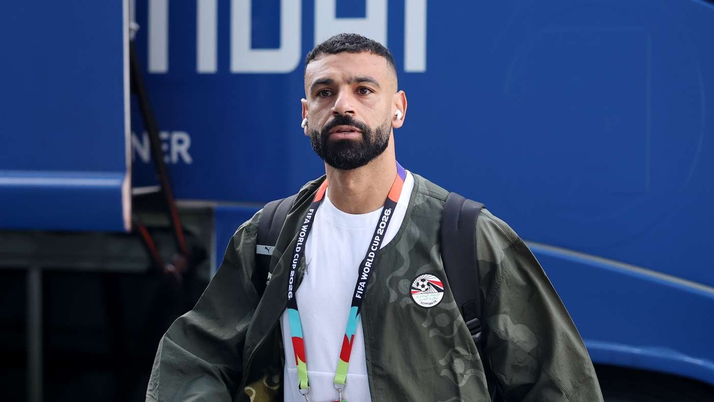 Man with a beard wearing a white shirt and camo jacket, headphones in, standing in front of a blue vehicle with a World Cup lanyard visible.