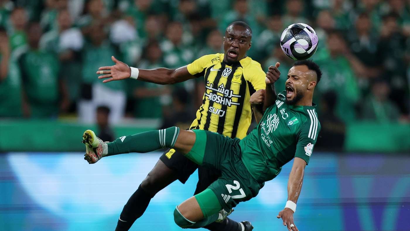 Two soccer players race for the ball in a high-speed contest; one in a yellow-and-black striped kit and the other in green, with a crowd in the background.
