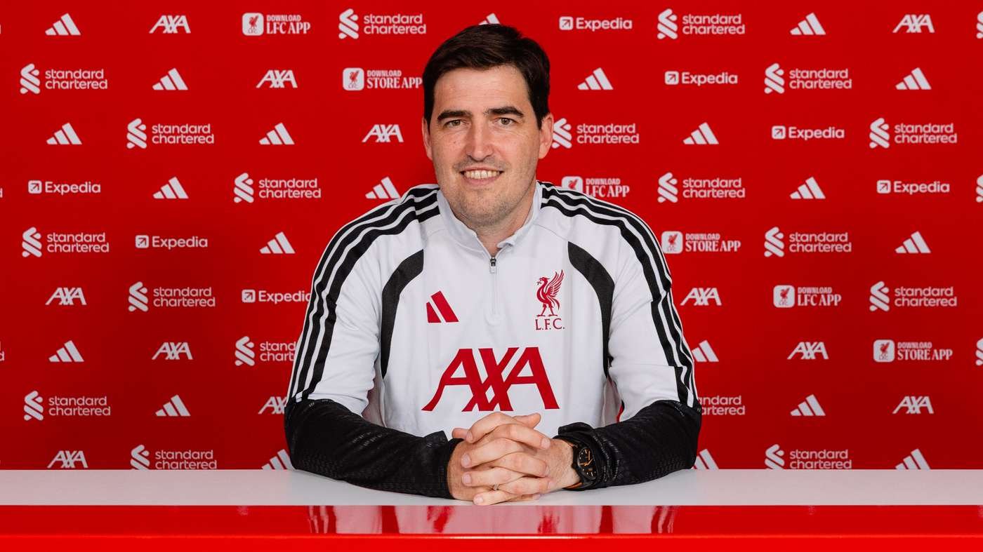 Man in a white Liverpool FC training top sits at a desk in front of a red sponsor backdrop with AXA logos and L.F.C. crest clearly visible. Man in a white Liverpool FC training top sits at a desk in front of a red sponsor backdrop with AXA logos and L.F.C. crest clearly visible.