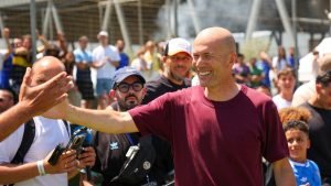 Bald man in a maroon shirt extends his arm to greet a crowd at an outdoor event.