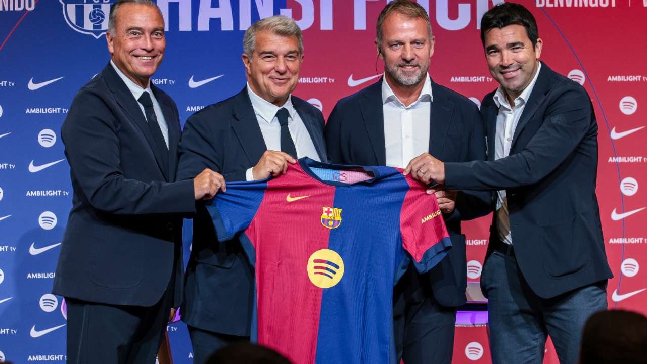Four men in suits pose on stage holding a blue-and-red FC Barcelona jersey during a press event or sponsorship announcement. Four men in suits pose on stage holding a blue-and-red FC Barcelona jersey during a press event or sponsorship announcement.