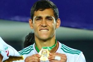 Male athlete proudly displaying a gold medal, wearing a white jersey with green trim on a podium. Male athlete proudly displaying a gold medal, wearing a white jersey with green trim on a podium.