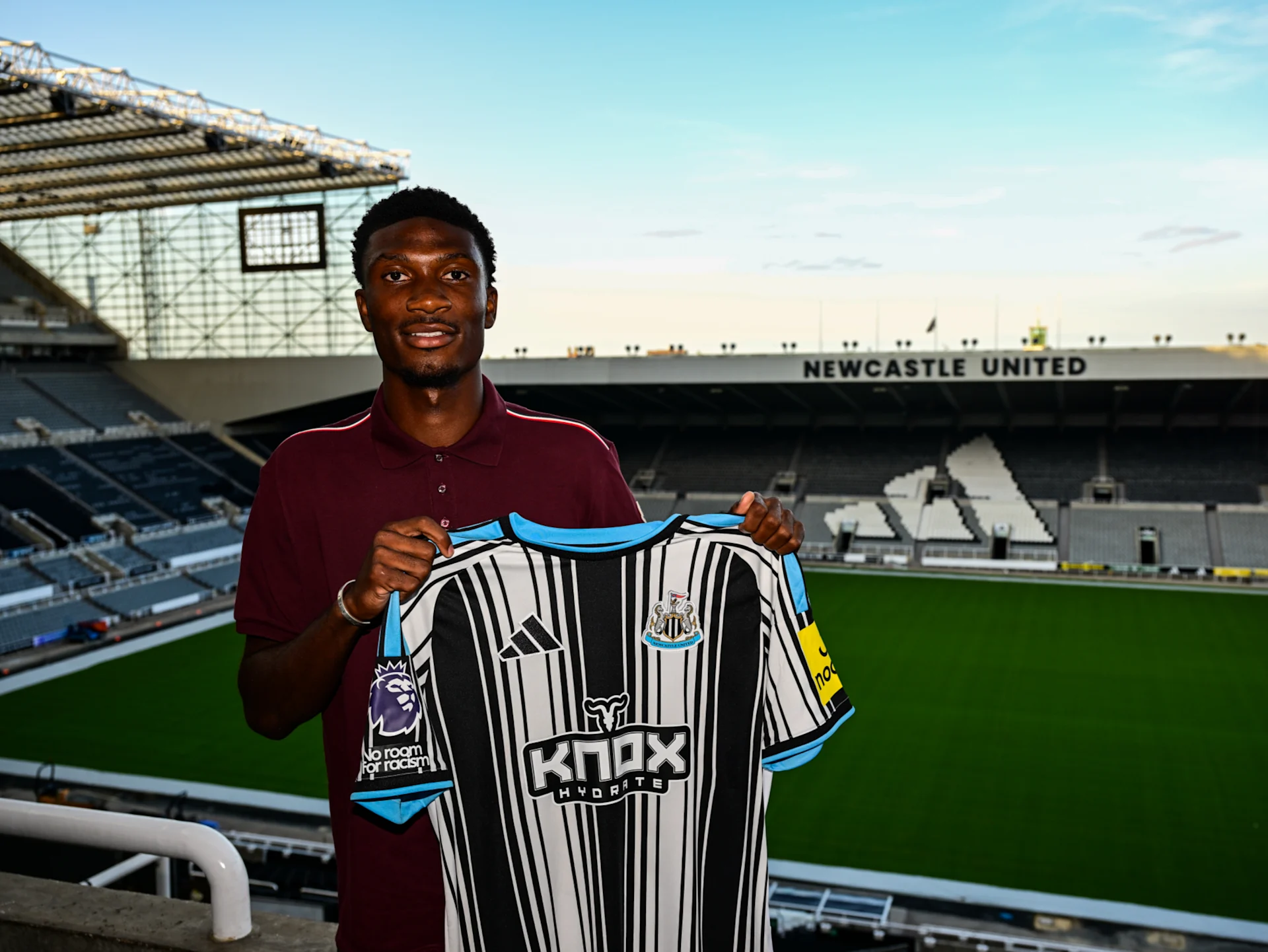 Young man standing in an empty soccer stadium, holding a Newcastle United jersey with a black-and-white stripe design in front of him. The pitch and stands are visible behind him. Young man standing in an empty soccer stadium, holding a Newcastle United jersey with a black-and-white stripe design in front of him. The pitch and stands are visible behind him.