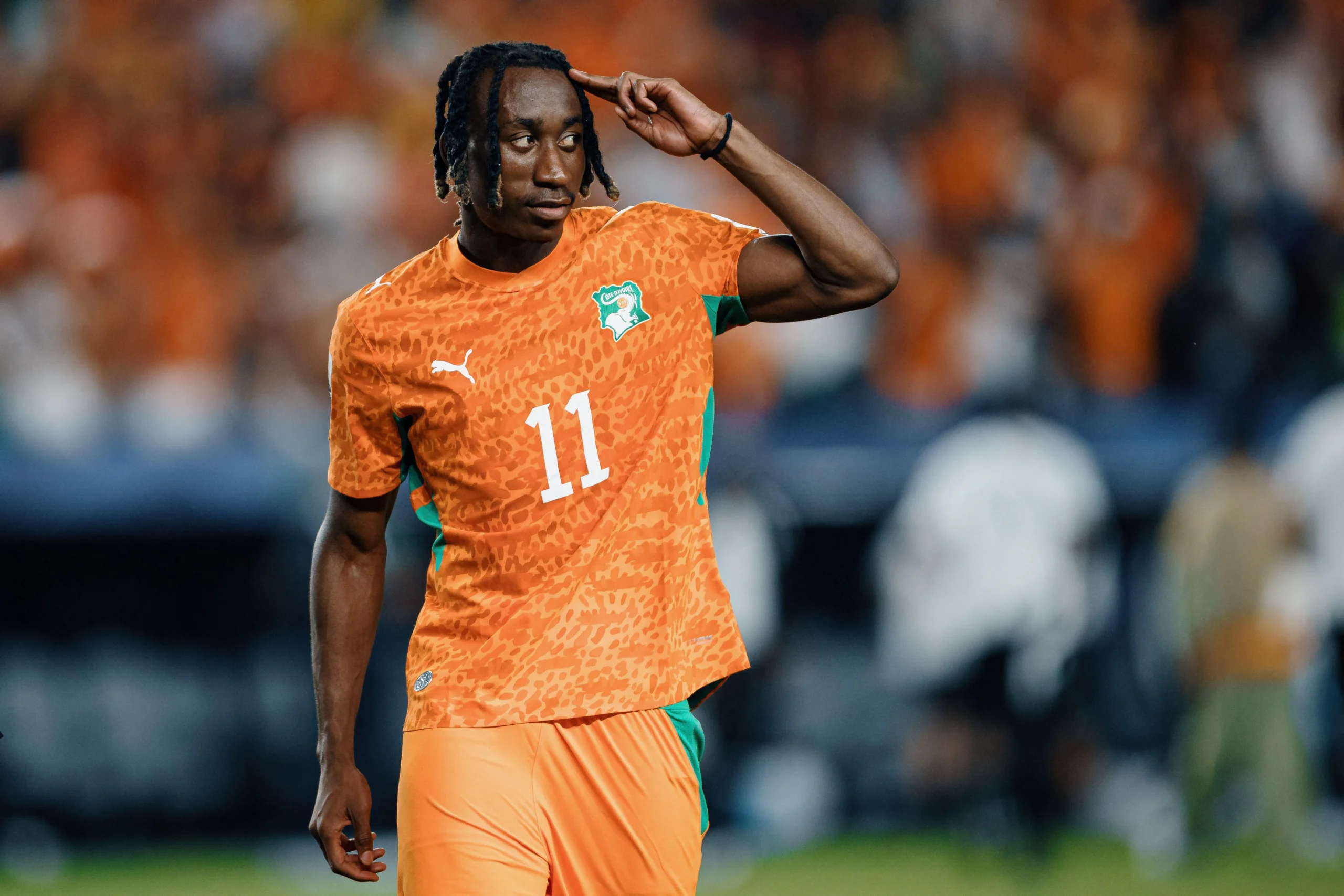 Soccer player in an orange kit (No. 11) salutes with his hand at his head on the field, blurred crowd behind him.