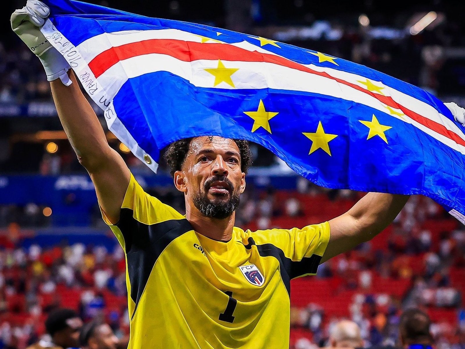 Male soccer player in a yellow jersey raises a blue flag with yellow stars over his head during a game in a stadium. Male soccer player in a yellow jersey raises a blue flag with yellow stars over his head during a game in a stadium.