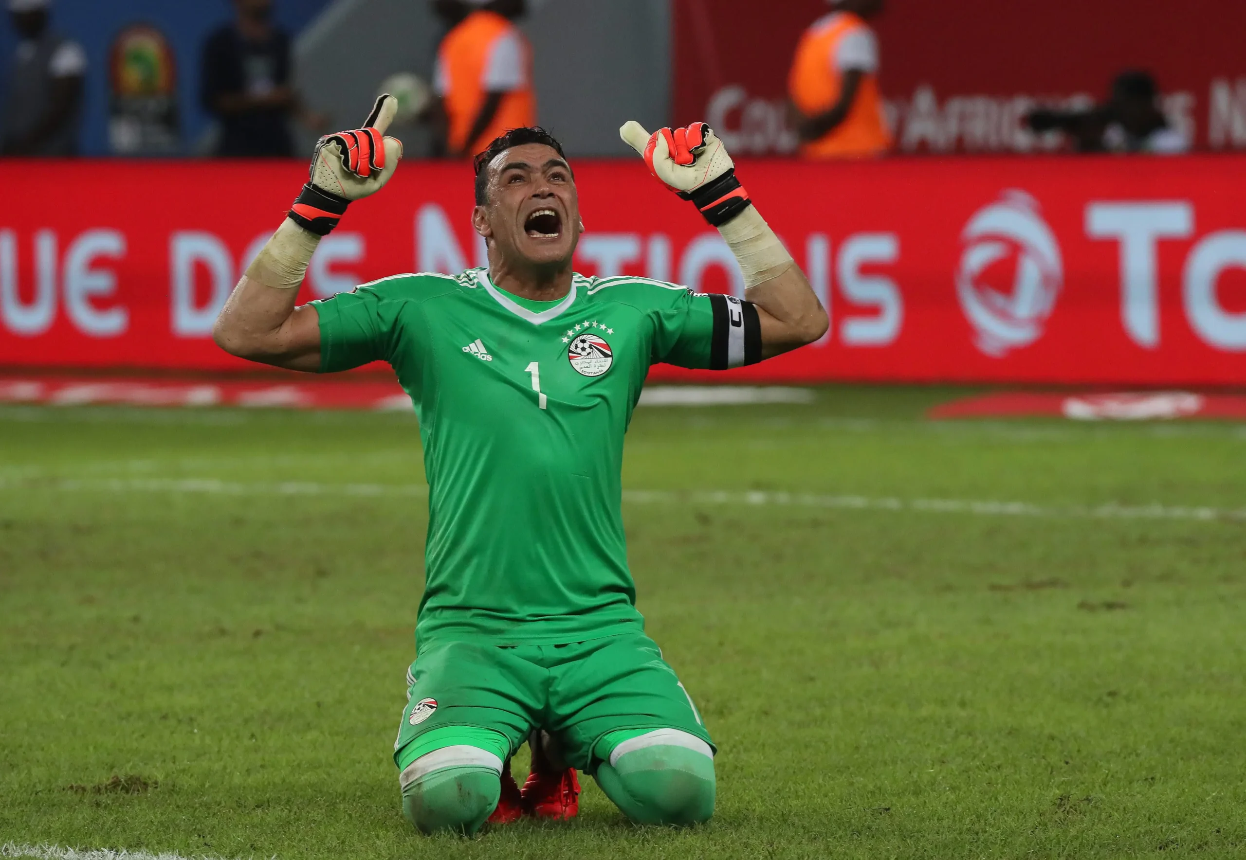 Egyptian national team goalkeeper in green, kneeling on the pitch with fists raised in celebration; red advertising boards behind him. Egyptian national team goalkeeper in green, kneeling on the pitch with fists raised in celebration; red advertising boards behind him.