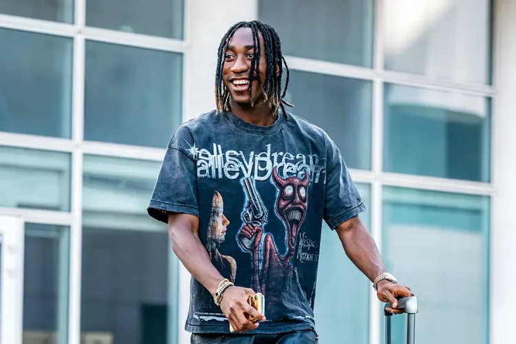 A smiling man with dreadlocks wearing a graphic t-shirt stands outside a glass building, holding a small suitcase handle and a phone. A smiling man with dreadlocks wearing a graphic t-shirt stands outside a glass building, holding a small suitcase handle and a phone.