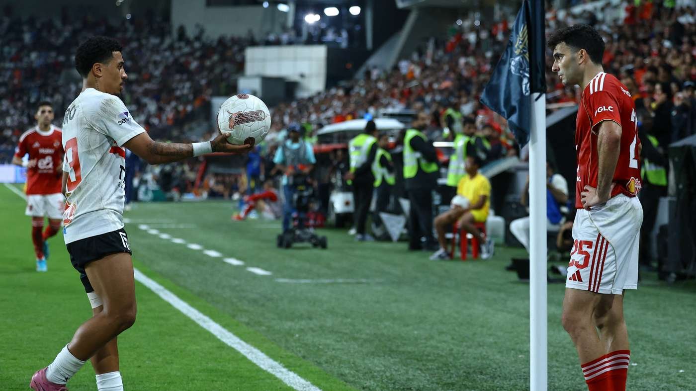 Soccer player in a white jersey prepares a throw-in by the sideline as a red-jersey opponent watches near the corner flag.