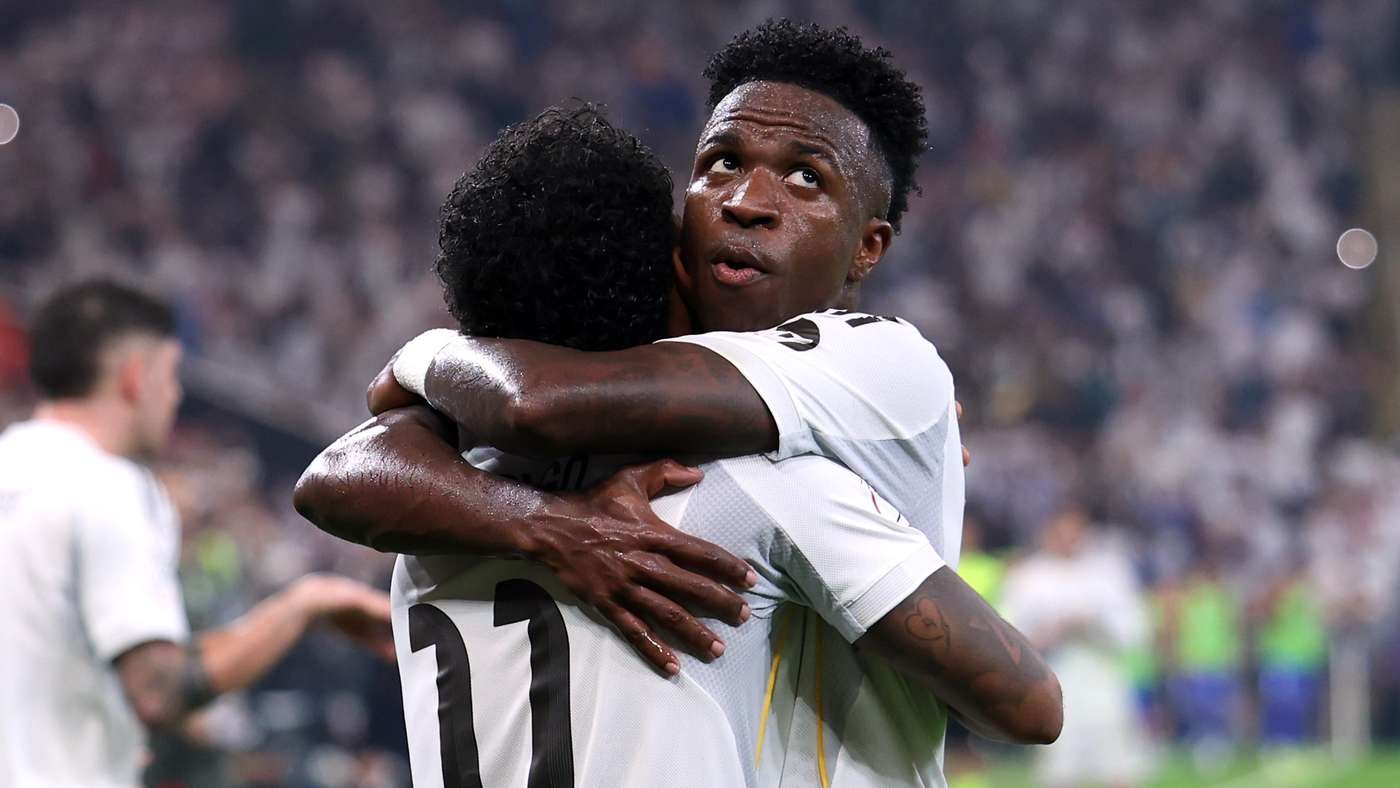 Two soccer players in white jerseys embrace in celebration on the field, stadium crowd blurred in the background.