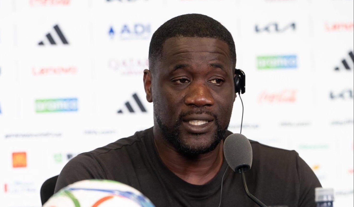 Man in a black shirt speaks into a microphone at a press conference, wearing a headset with logos blurred in the background. Man in a black shirt speaks into a microphone at a press conference, wearing a headset with logos blurred in the background.