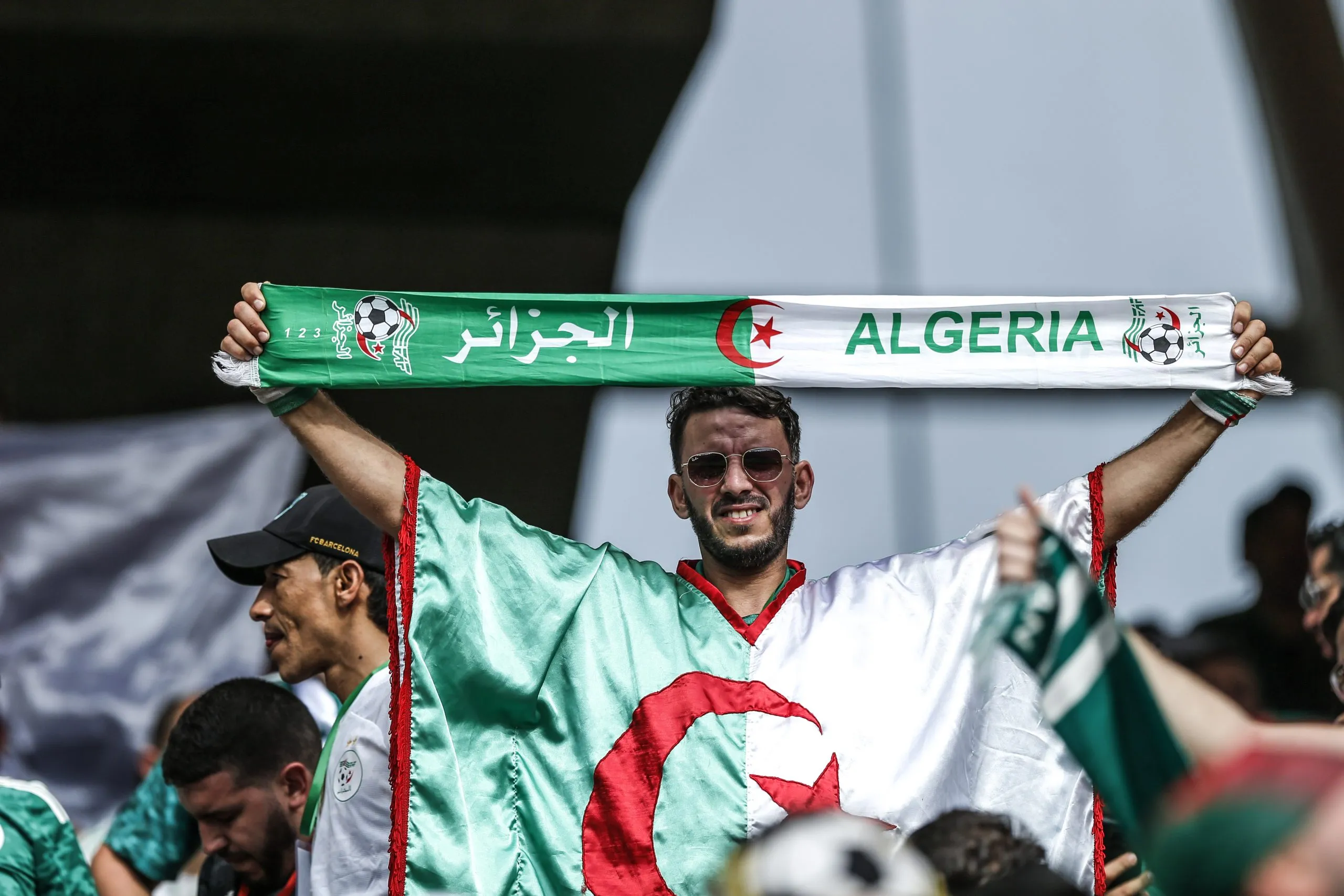 Algeria football fans hold a green-and-white scarf with 'ALGERIA' and a red star and crescent logo, celebrating in stadium stands. Algeria football fans hold a green-and-white scarf with 'ALGERIA' and a red star and crescent logo, celebrating in stadium stands.