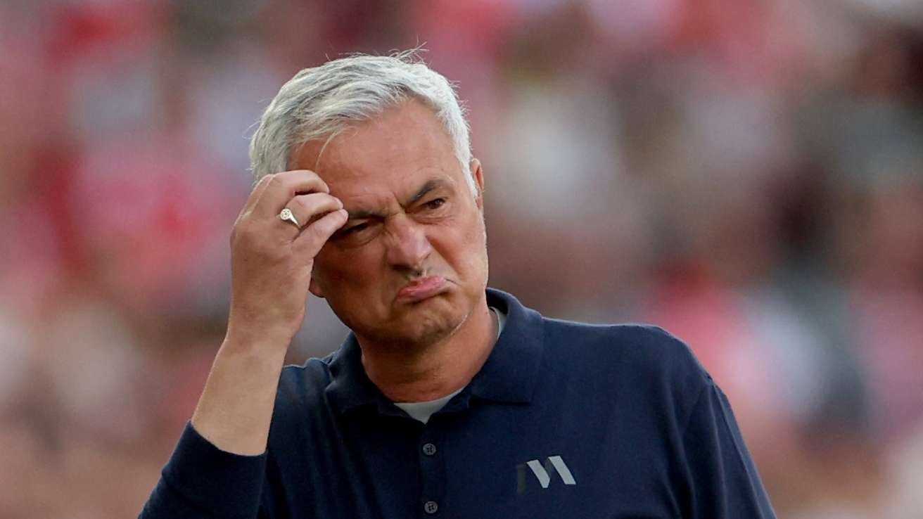 Man with gray hair in a navy polo scrunches his face and touches his temple, as if puzzled, in a blurred outdoor stadium. Man with gray hair in a navy polo scrunches his face and touches his temple, as if puzzled, in a blurred outdoor stadium.