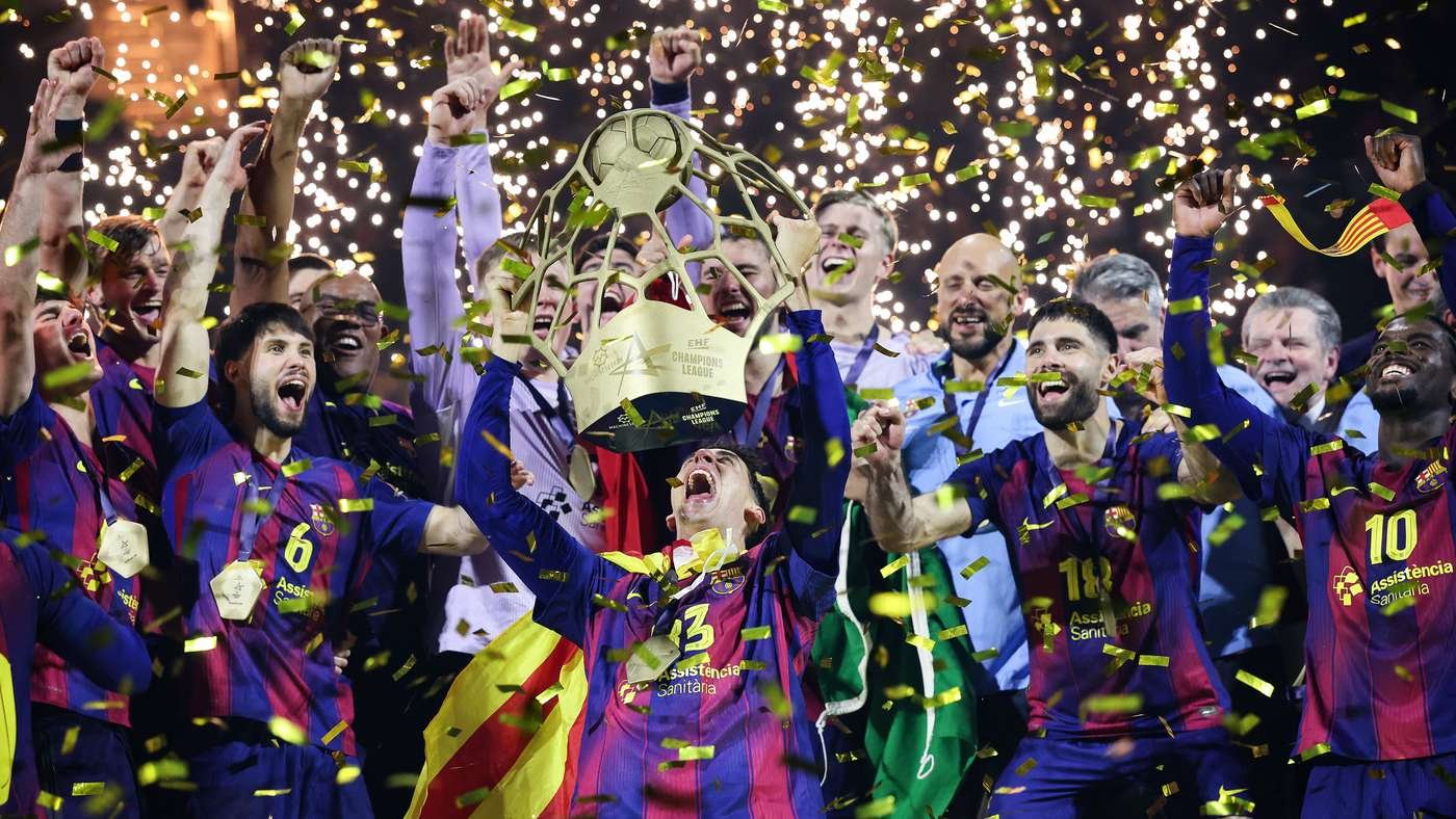 Soccer players in purple and red uniforms celebrate victory, lifting a large trophy amid gold confetti and cheering fans.