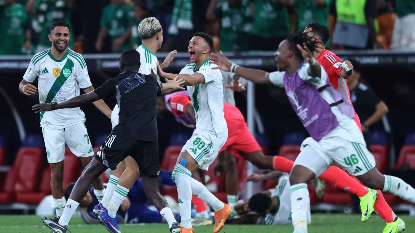 Soccer players in white and green celebrate on the field, embracing and smiling as fans react in the background.