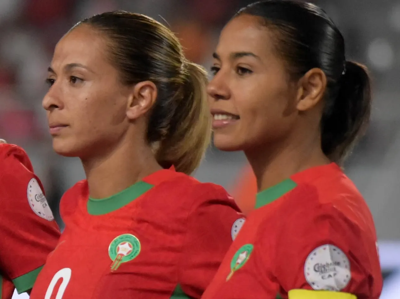 Two female soccer players in red jerseys with green trim standing side by side on the field, looking ahead.