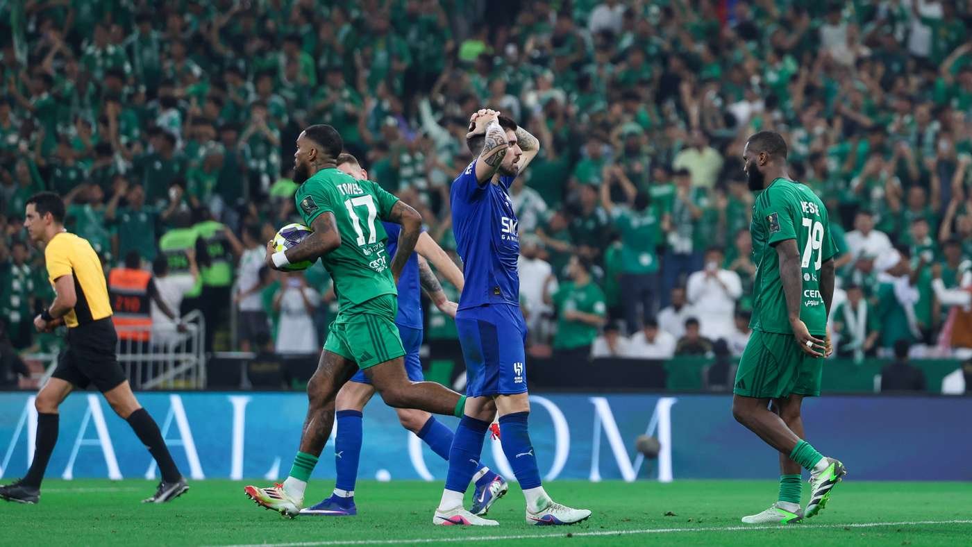 Soccer players on the field: a green team member holds a ball as a blue-clad opponent raises his arms, with a yellow referee nearby and a packed green crowd in the stands behind them.