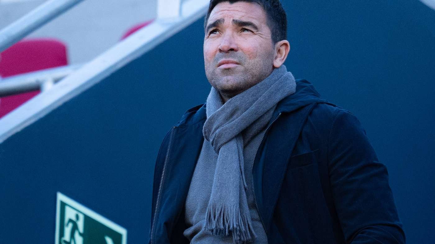 Man in a gray scarf and dark jacket gazing upward, standing beside blue walls outdoors, pensively.