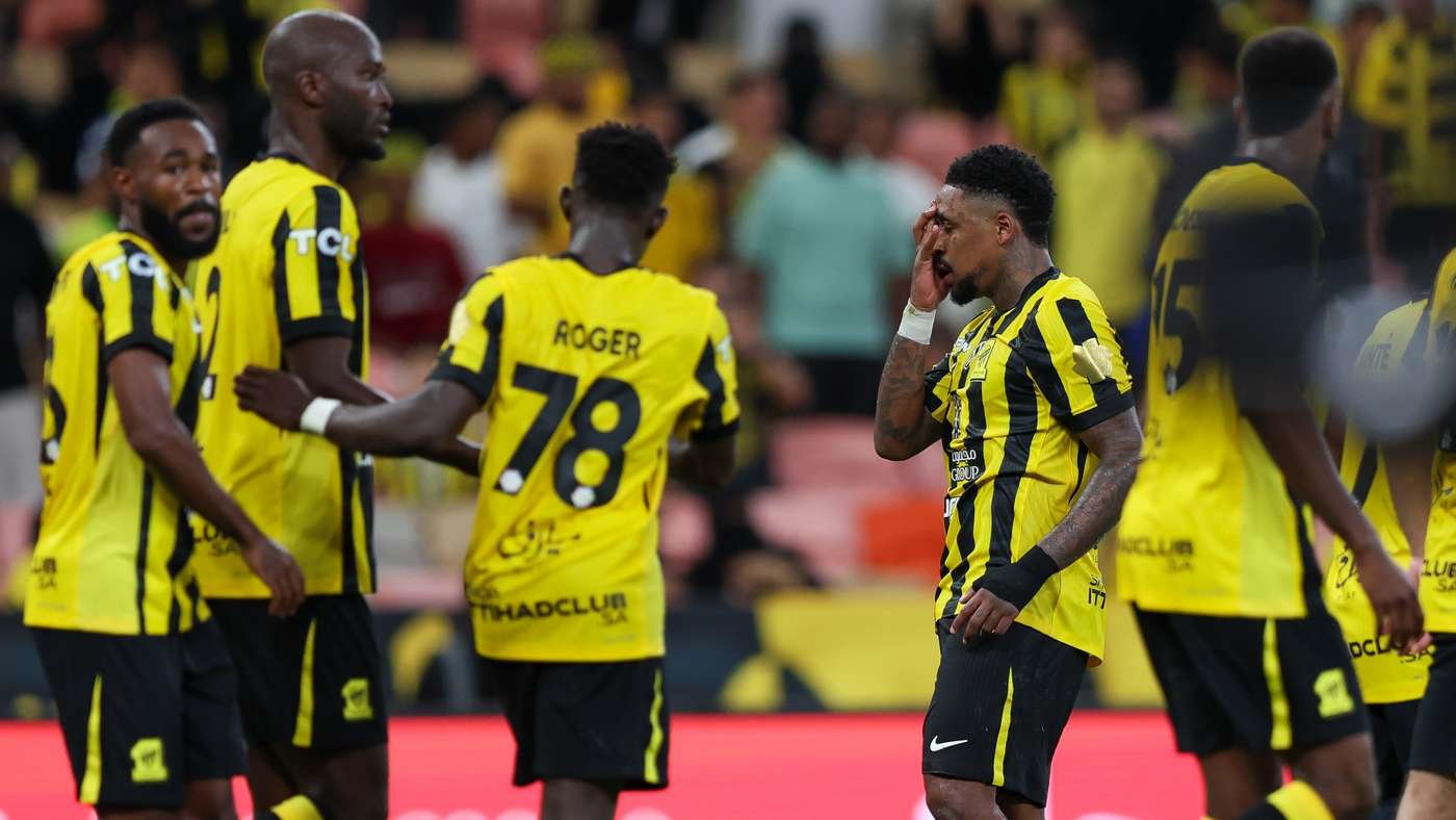 Soccer players in yellow and black striped uniforms standing and walking on a field, crowds in the stands behind them.