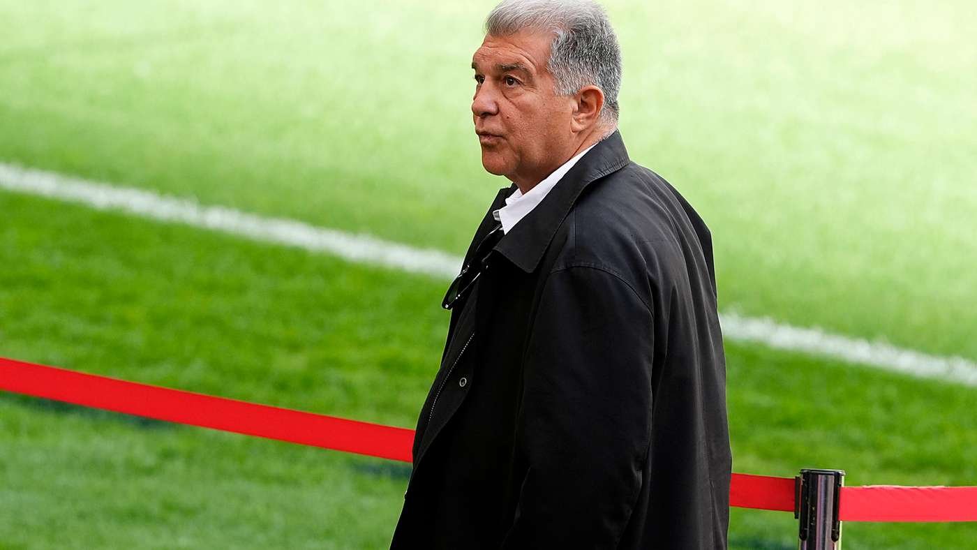 A man in a black coat stands on a grassy sports field beside a red barrier, looking to the left.