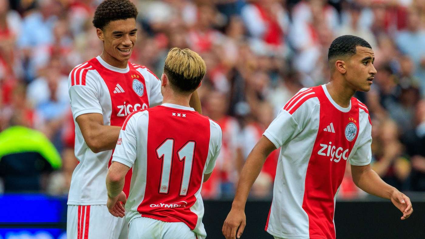Three Ajax players in red-and-white kits celebrate on the pitch after a goal, with a cheering crowd in the background.