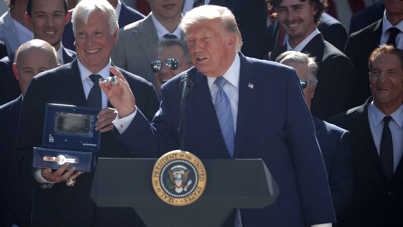 Man in a dark suit speaks at a lectern with the presidential seal, holding a medal and an open blue display case; group of suited men behind him.