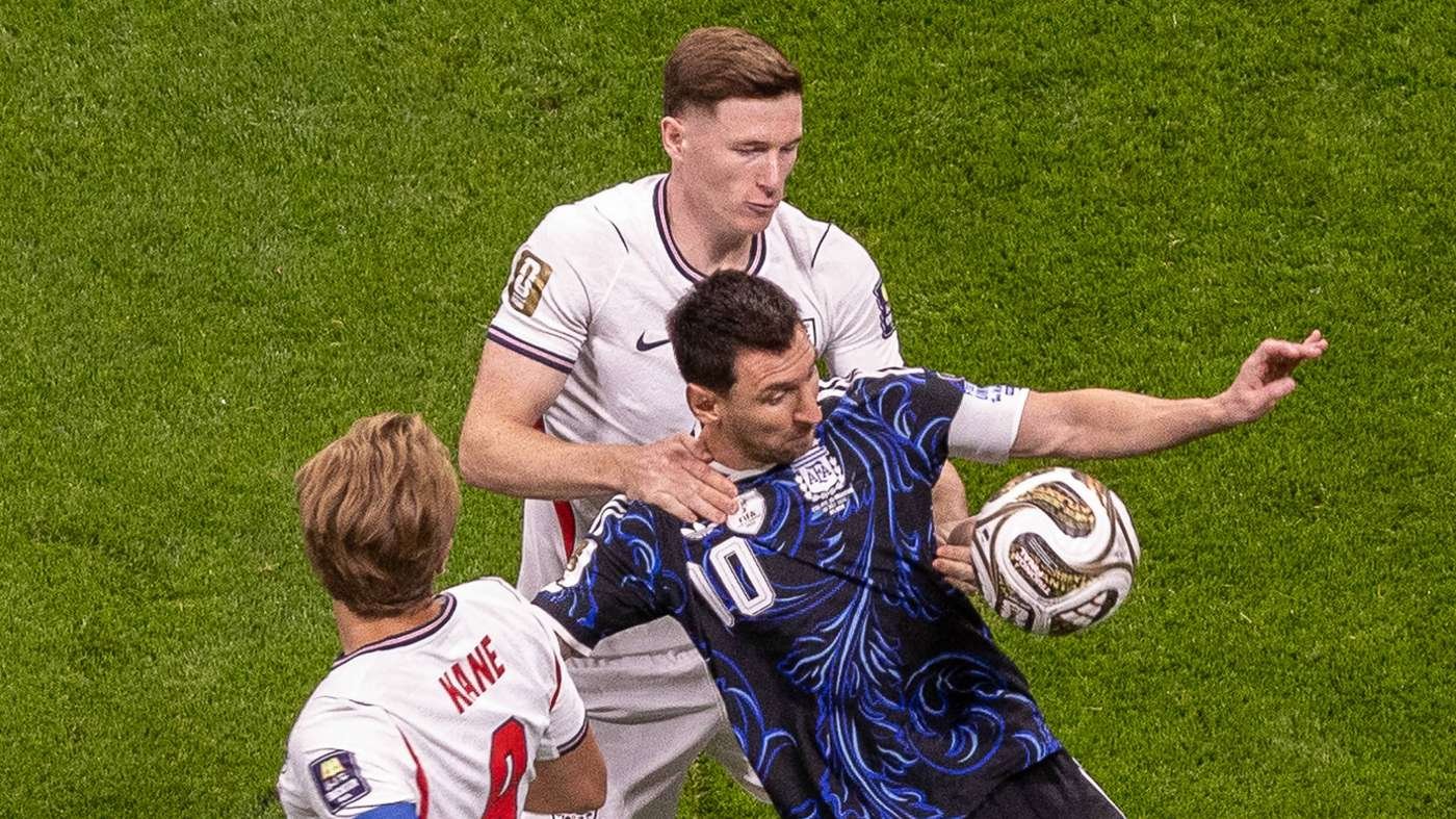 Three soccer players contest the ball on a grassy field, two in white jerseys closing in on a blue-patterned opponent.