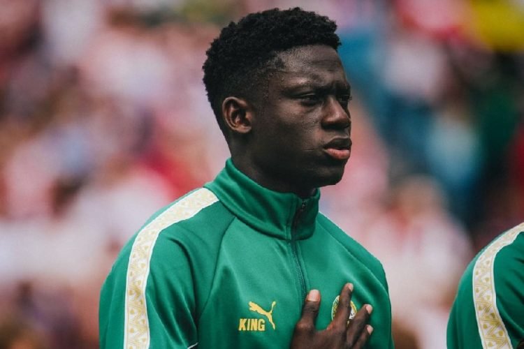Young Black male athlete in a green tracksuit with gold Puma logo and a 'KING' badge, hand over heart against a blurred stadium backdrop