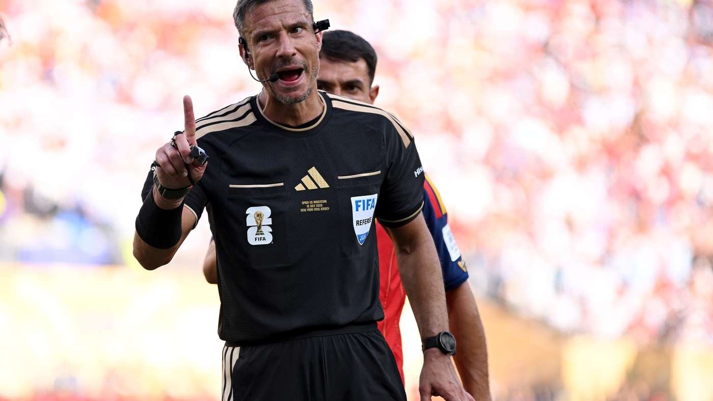 Soccer referee on the pitch raises one finger, wearing a black kit with a FIFA badge and headset, crowd blurred in the background.