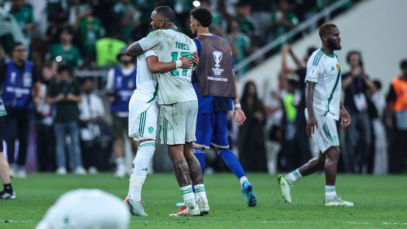 Two soccer players in white and green jerseys hug on a grass field, celebrating during a match with fans in the stands nearby.