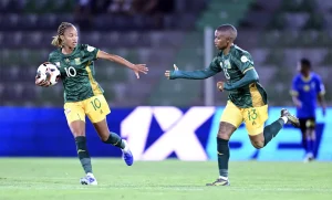 Two female soccer players in green jerseys chase the ball on the field during a match, one wearing number 10 with the ball at her feet.