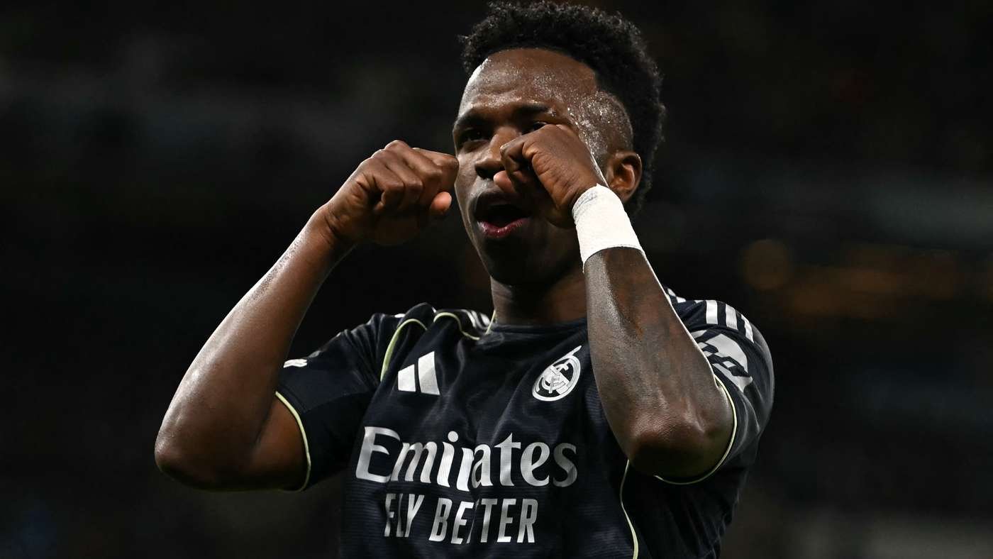 Soccer player in a dark kit celebrating a goal with fists raised near his face on a dim stadium backdrop.
