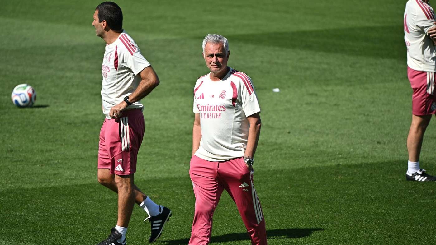 Soccer players in pink shorts and white shirts practice on a green field; the central man with gray hair faces the camera while others stand nearby.