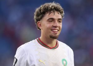 Male soccer player in a white jersey with a green crest, smiling on the field.
