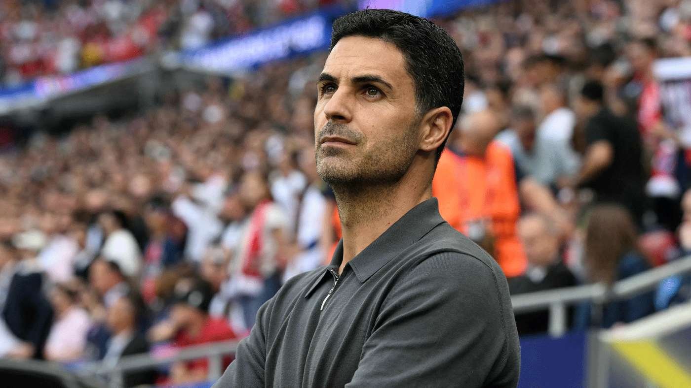 Man with short dark hair and stubble wearing a gray polo, standing with arms crossed as a crowded stadium fills the background, looking serious. Man with short dark hair and stubble wearing a gray polo, standing with arms crossed as a crowded stadium fills the background, looking serious.