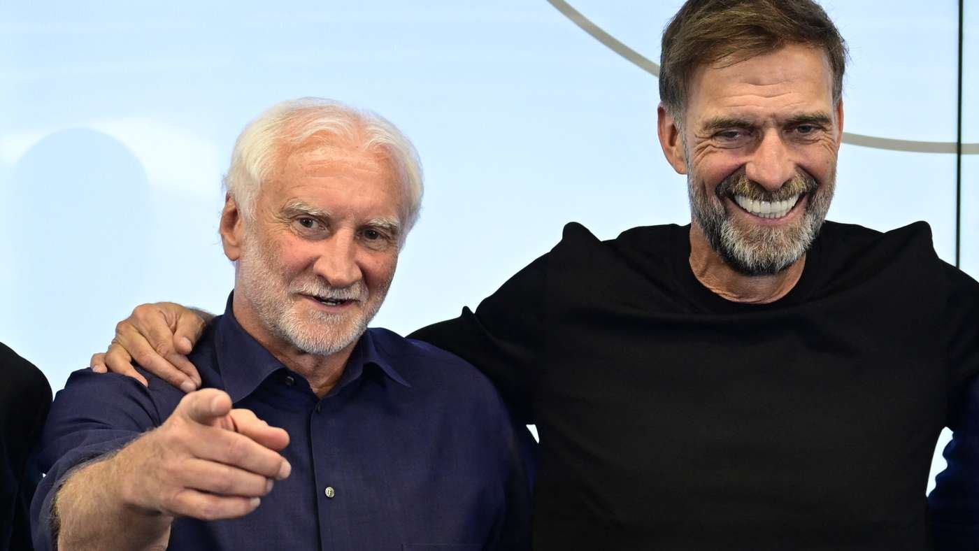 Two smiling men pose together, one pointing toward the camera with a hand on the other's shoulder against a light blue background. Two smiling men pose together, one pointing toward the camera with a hand on the other's shoulder against a light blue background.