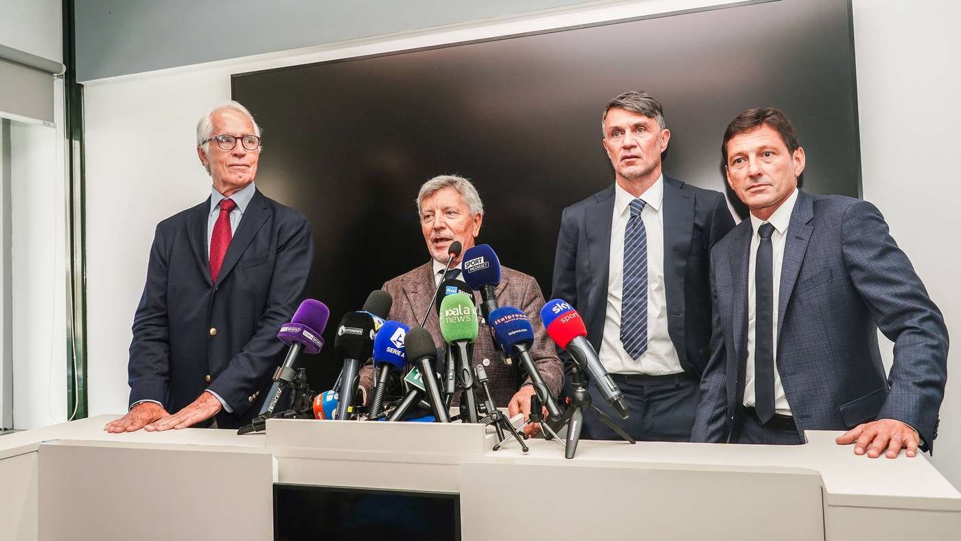 Four men in suits stand behind a white podium with numerous microphones at a press conference. Four men in suits stand behind a white podium with numerous microphones at a press conference.