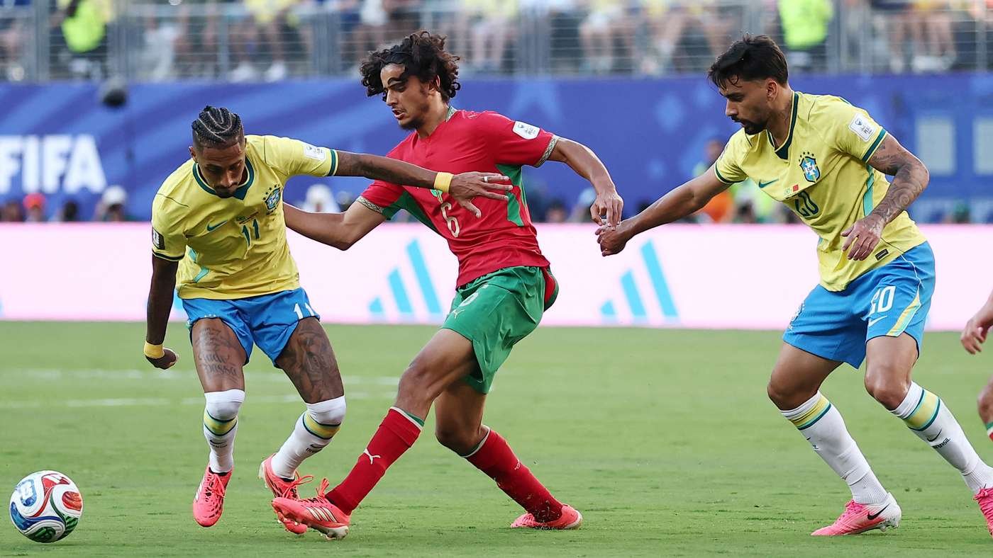 Three players clash for the ball in a soccer match: two Brazilians in yellow shirts chase a red‑green opponent on a green pitch. Three players clash for the ball in a soccer match: two Brazilians in yellow shirts chase a red‑green opponent on a green pitch.