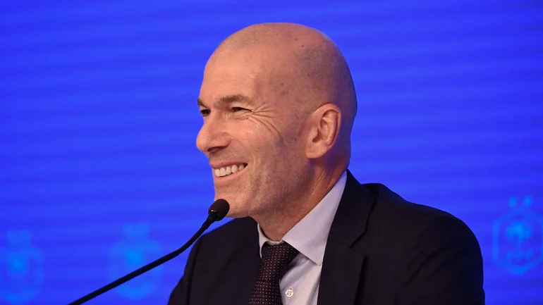Bald man in a dark suit smiling while speaking into a microphone at a conference with a blue backdrop
