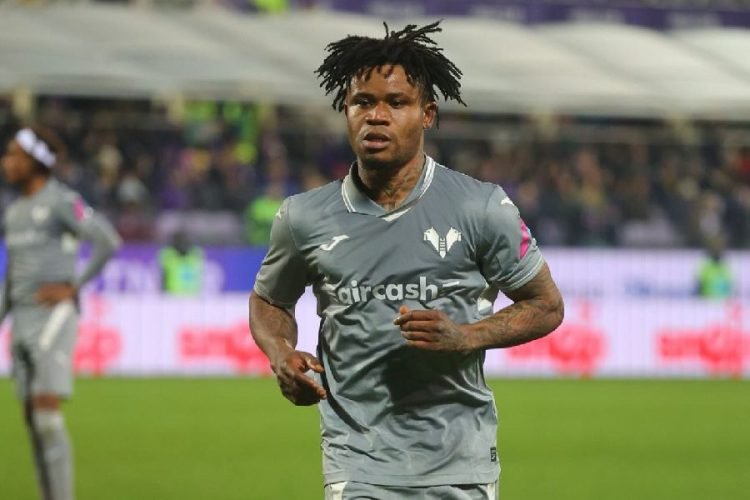 Soccer player in gray kit running on a pitch during a match, with a stadium in the background.
