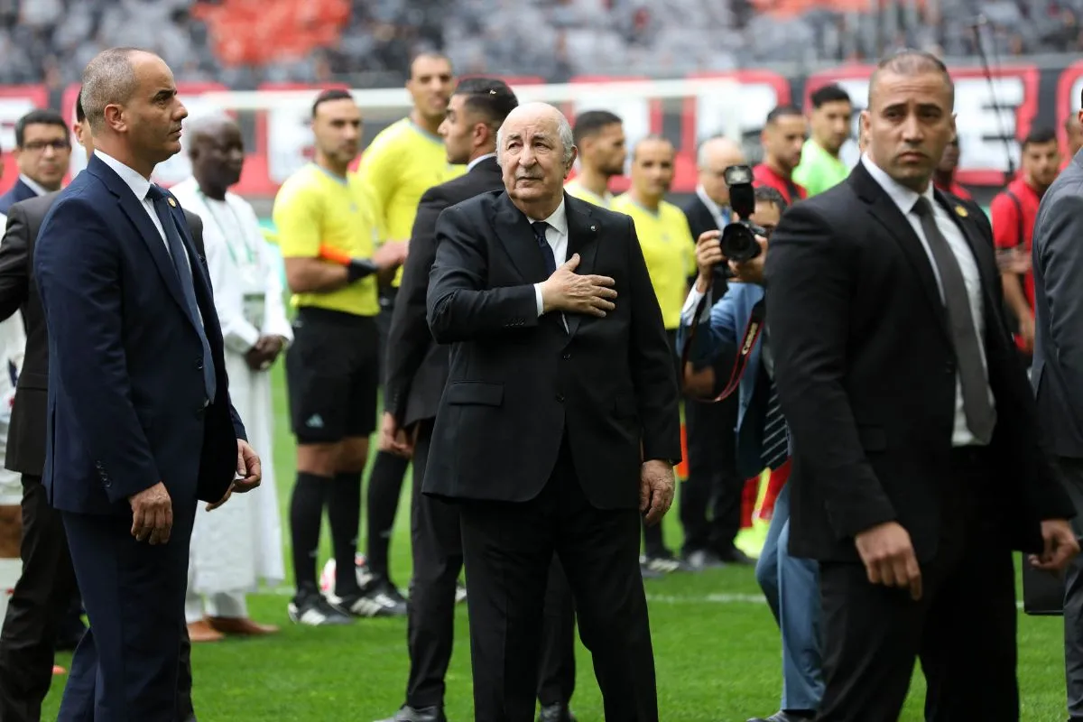 Older man in a dark suit places his hand over his heart on a soccer field, surrounded by photographers and security during a formal event.