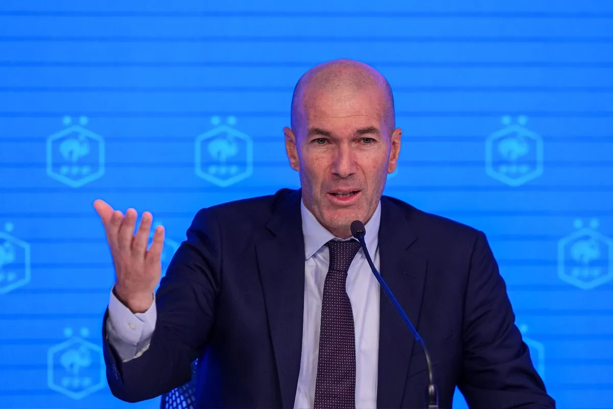 Bald man in a dark suit speaks into a microphone at a press conference, gesturing with his left hand against a blue backdrop with logos/branding.