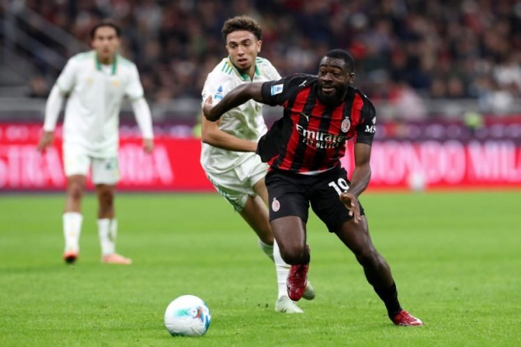 AC Milan forward in a black and red kit dribbles the ball as a defender in white-green chases from behind on a soccer pitch.