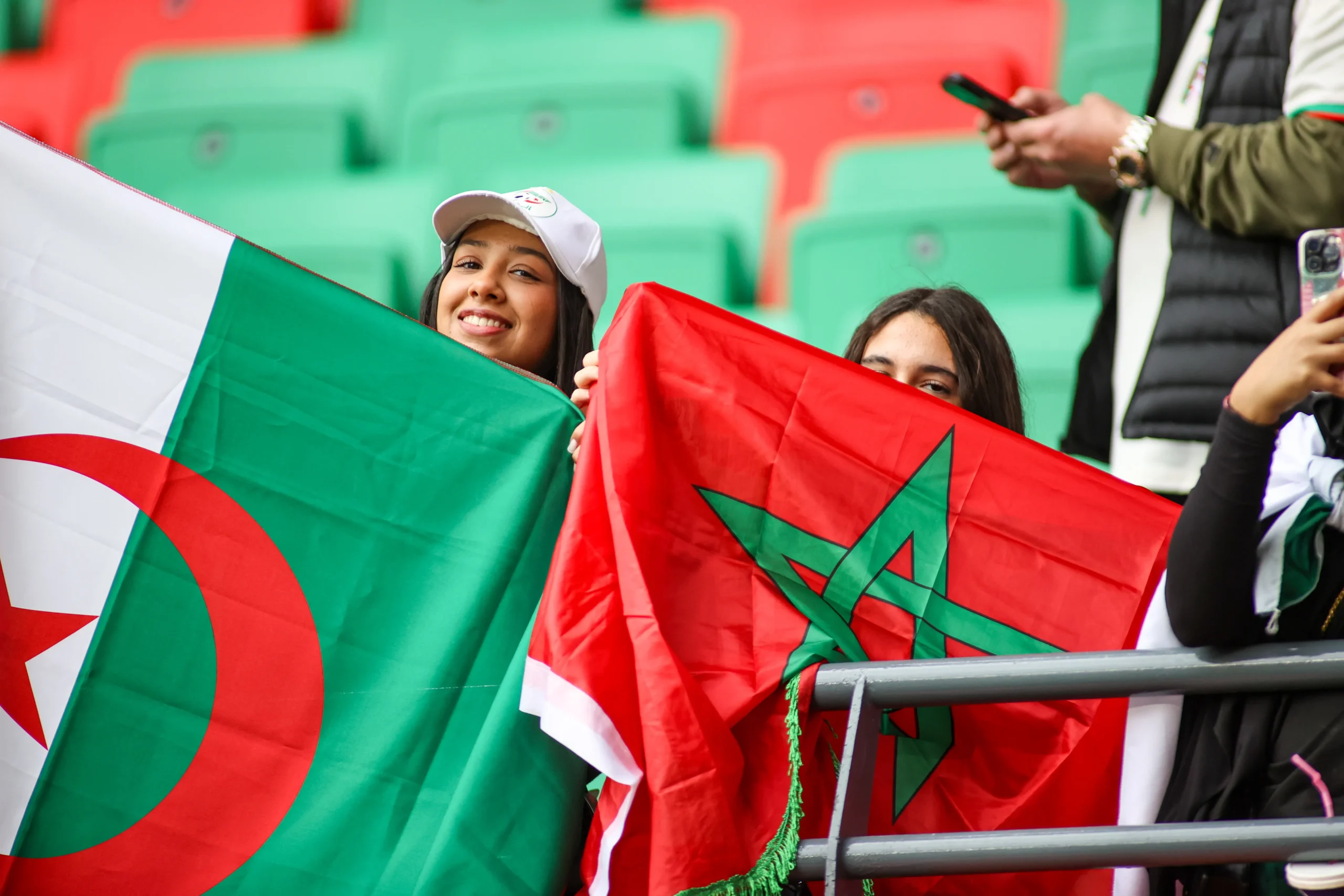 Smiling fan wearing a white cap, waving a green-and-red flag at a stadium while another person holds a red flag with a green star in the foreground.