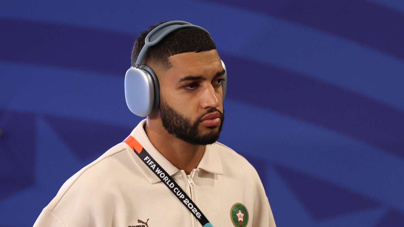 Man wearing large gray over-ear headphones and a beige polo, with a FIFA World Cup 2026 lanyard, against a blue striped background.