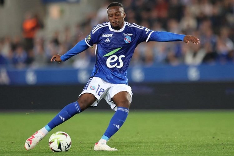 Soccer player in a blue kit kicking a white ball on a green field during a match, stadium blurred in the background. Soccer player in a blue kit kicking a white ball on a green field during a match, stadium blurred in the background.