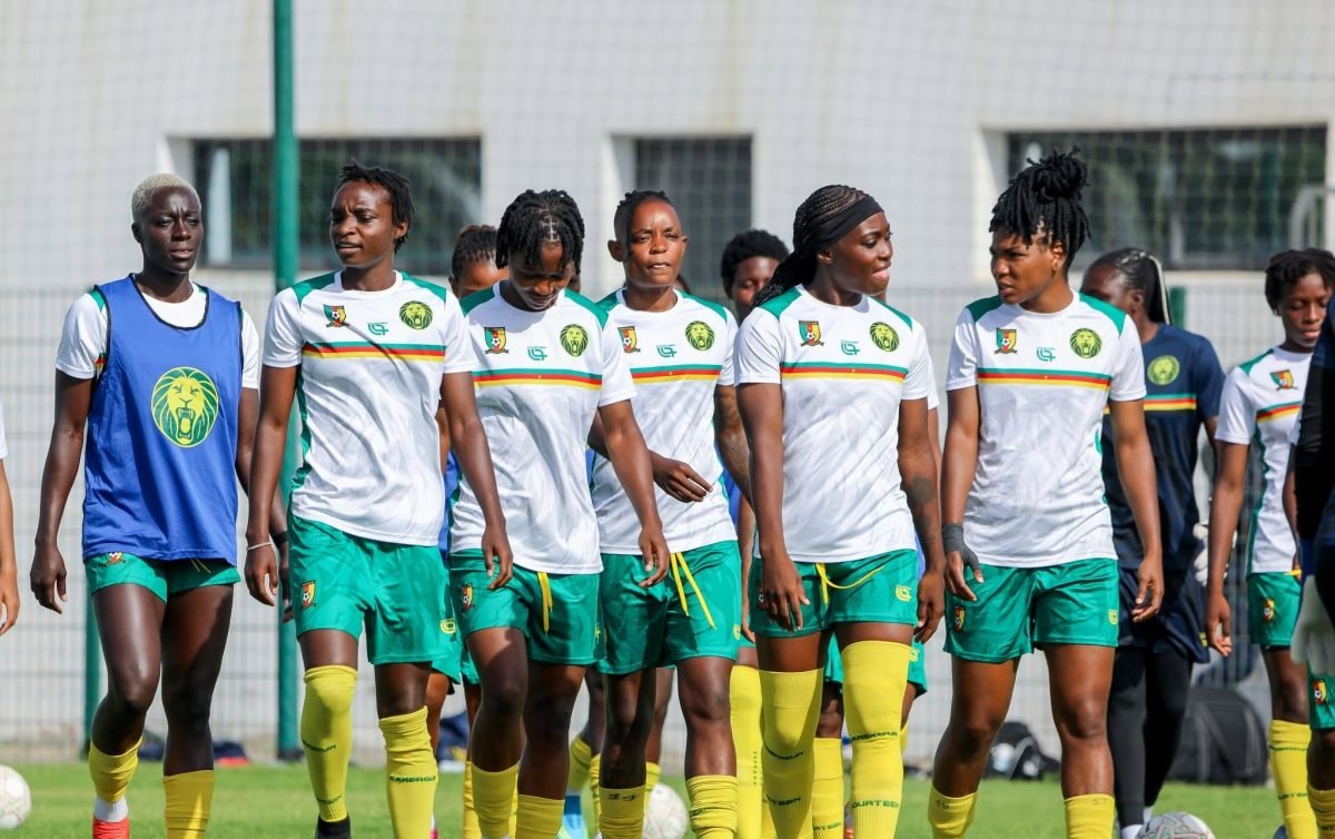 Group of female soccer players in white and green uniforms walking on a practice field, yellow socks visible, one in a blue bib on the left side.